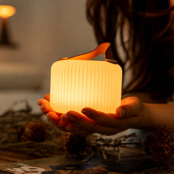 A person holds a glowing ribbed cylindrical lamp with a warm orange-brown handle in a dim indoor setting. The lamp emits a soft warm light, and blurred dried botanicals sit on a table in the foreground.