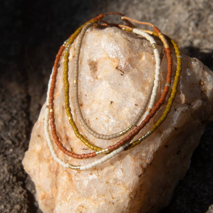 Three beaded bracelets in rust orange, olive green, and cream white, coiled on a pale pinkish stone against a dark textured rock background.