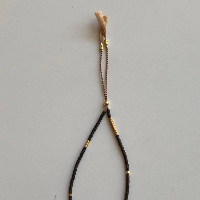 A black braided cord bracelet with small gold metallic beads and a light tan tassel, displayed on a neutral light gray background.