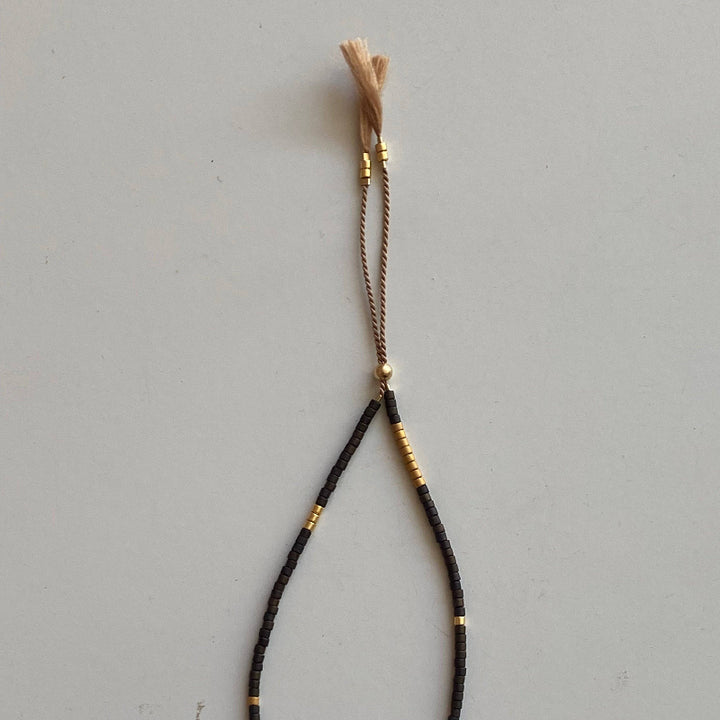 A black braided cord bracelet with small gold metallic beads and a light tan tassel, displayed on a neutral light gray background.