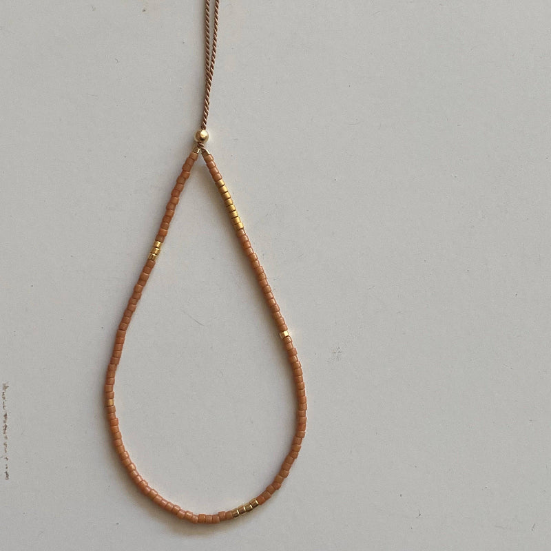 A delicate teardrop-shaped beaded necklace in a warm peachy-brown color with small gold metallic accents, featuring a matching tassel on a thin cord, displayed against a light gray background.