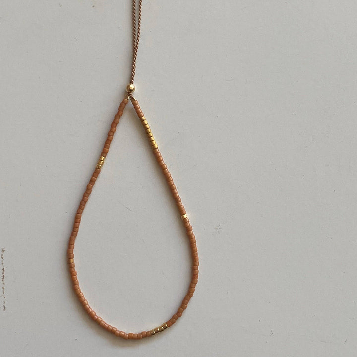 A delicate teardrop-shaped beaded necklace in a warm peachy-brown color with small gold metallic accents, featuring a matching tassel on a thin cord, displayed against a light gray background.