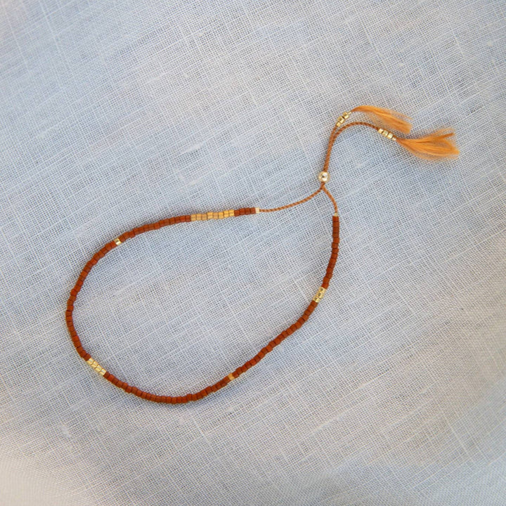 A delicate beaded necklace with a reddish-brown cord, small gold spacer beads, and a matching peach tassel, laid flat on a light blue textured fabric background.