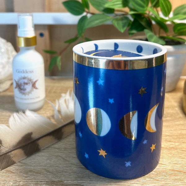 A blue ceramic candle holder with a gold-rimmed rim and a moon phase pattern, styled on a wooden tray next to a Goddess brand diffuser bottle and a green plant.