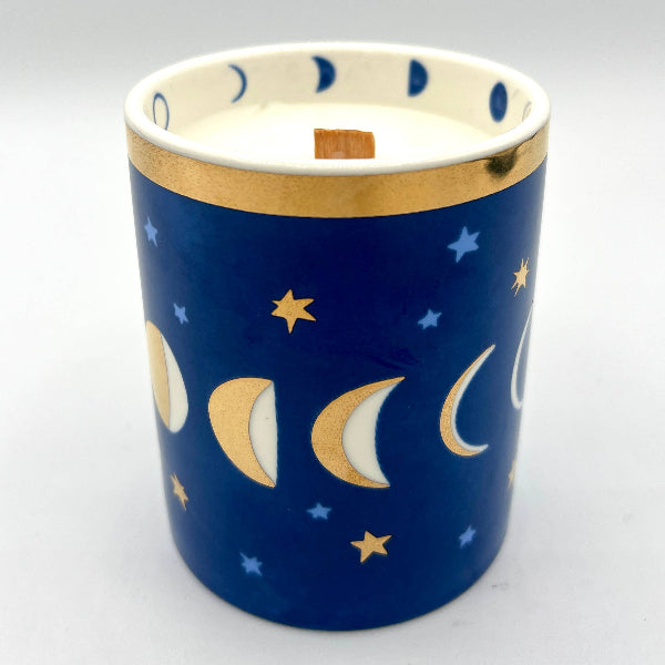 A ceramic candle holder with a deep blue body decorated with gold and white moon phases and stars, featuring a white interior and a gold band around the rim.