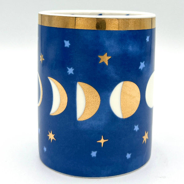A cylindrical ceramic candle holder with a deep blue body decorated with gold and white moon phases and stars, featuring a gold band around the rim.