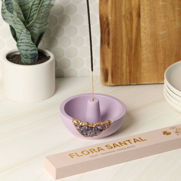 Purple incense holder with a lit incense stick on a white surface, next to a plant and a box of Flora Santal incense.